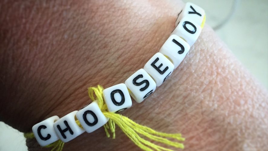 A close up of a bracelet made from beads that spell 'Choose Joy'. It is tied around a wrist with yellow string.